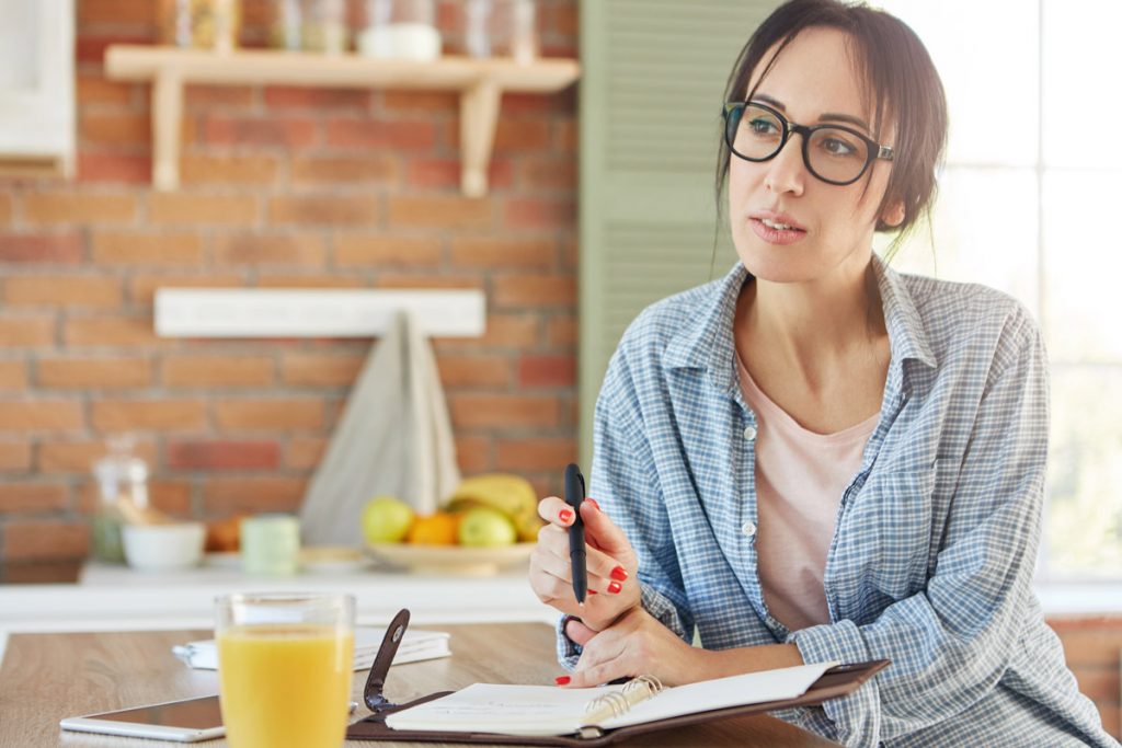 Young good looking woman wants to loose weight, writes diet plan in notebooks as sits on kitchen and drinks juice.