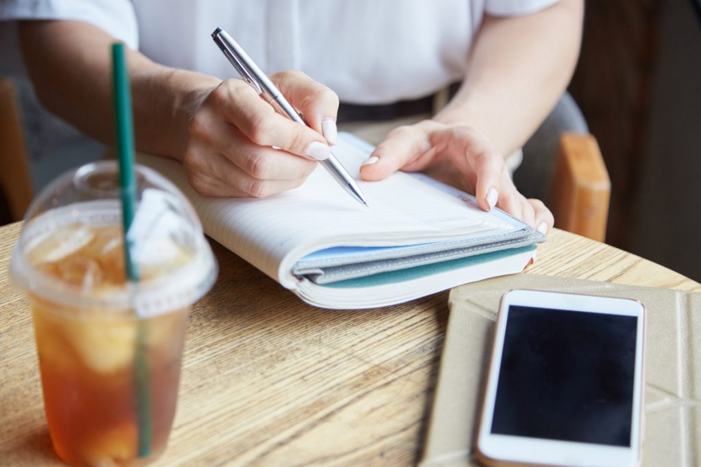 Hands of female student sitting at cafe table and writing essay in textbook.