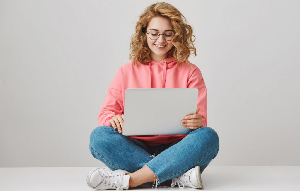 Happy attractive caucasian student with curly hair sitting on floor with crossed legs and holding laptop, typing or browsing network while wearing eyeglasses.