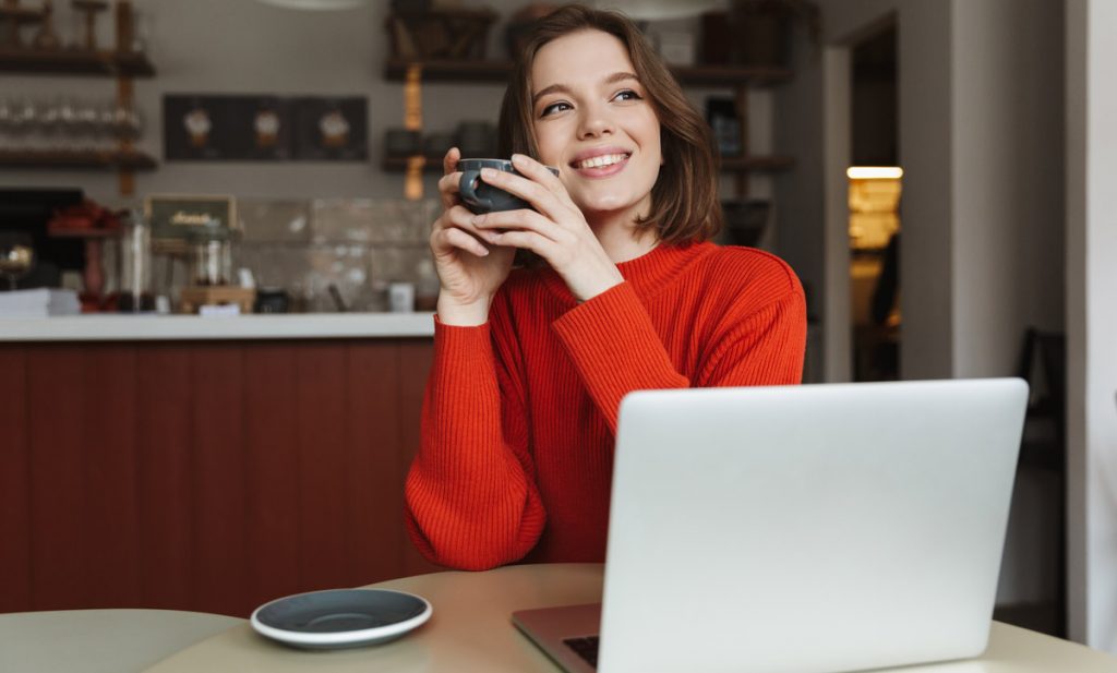 Image of happy caucasian woman 20s smiling while using laptop and drinking coffee in cafe.