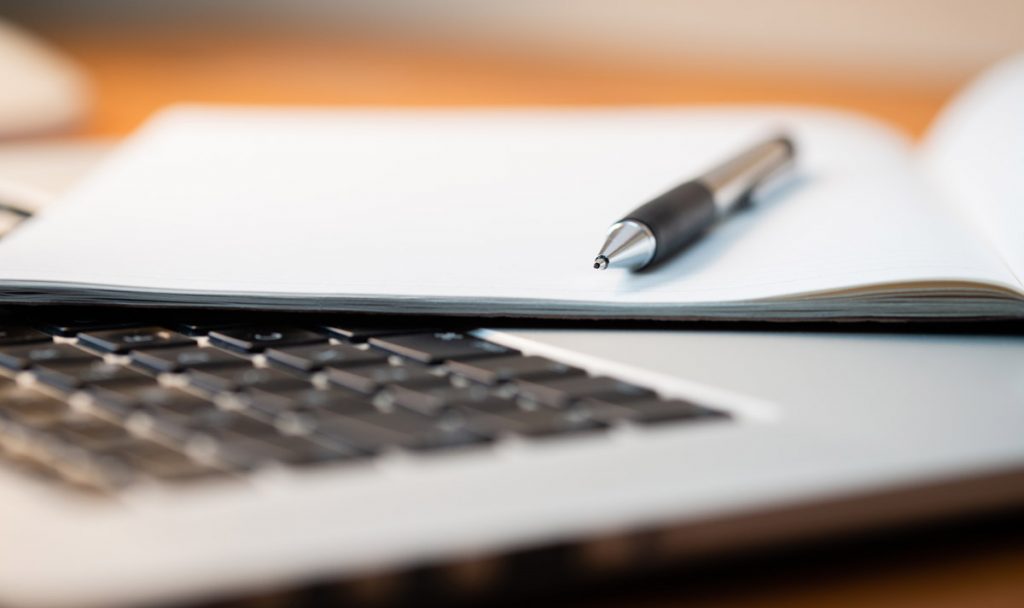 Laptop Computer on a Modern Wooden Business Desk with a Notepad and Pen in Unfocused Background.