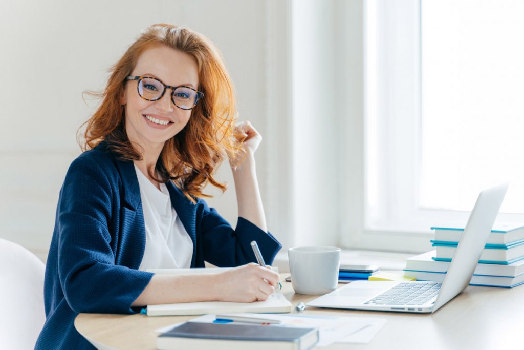 Sideways shot of prosperous businesswoman with foxy hair, smiles positively, records information in notepad, drinks coffee, searches information on laptop computer, develops strartup project.